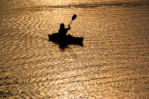 Silhouette of one person in a kayak paddling on a rippling river at sunset.