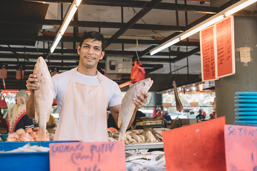 a fish stall owner holding fishes on both of his hand looking at camera smiling in front of his stall