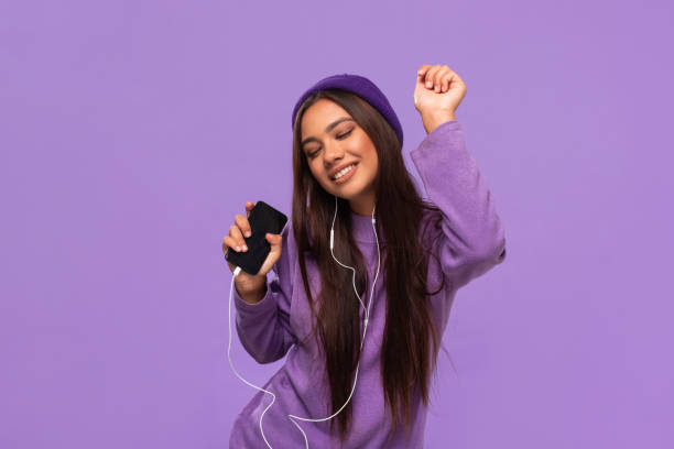 Pretty african-american girl in a hat and sweater listening to music on a mobile phone with wired headphones and dancing isolated over purple background. Enjoying life. Pretty african-american girl in a hat and sweater listening to music on a mobile phone with wired headphones and dancing isolated over purple background. Enjoying life. headphones plugged in photos stock pictures, royalty-free photos & images
