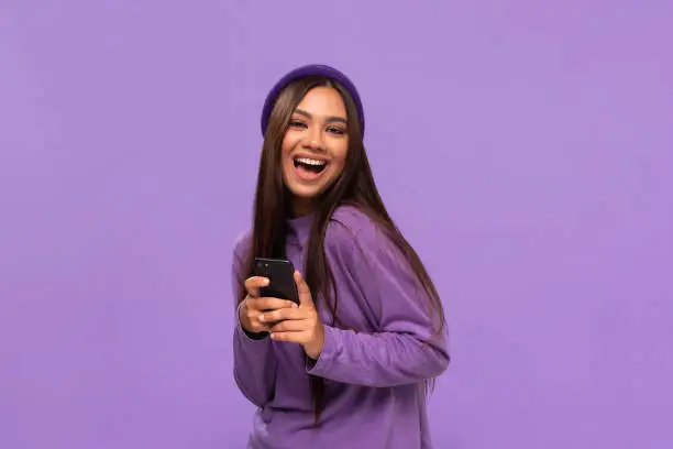 Photo of Smiling african-american girl in a hat and sweater holding smartphone standing isolated over purple background.