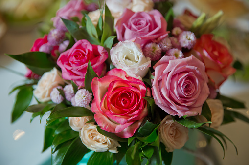 Closeup photo of a beautiful bouquet of pastel cream and pink roses, yellow and pink honeysuckle and Hawthorne blossom in a glass vase on a rustic wooden table in front of a pale curtain