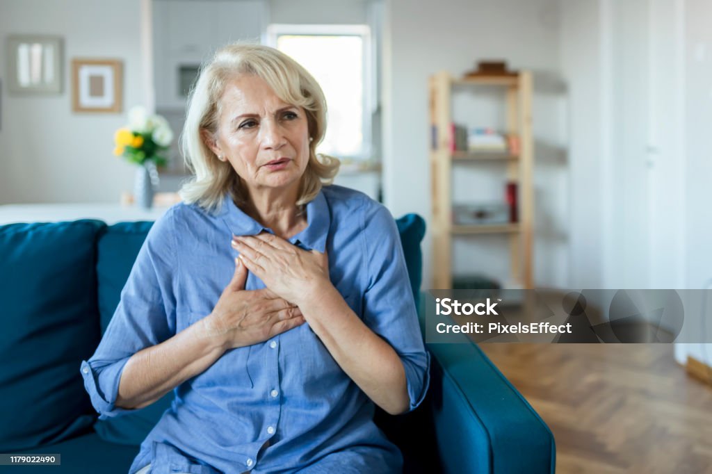 Upset stressed older woman feeling heartache Senior Woman Suffering From Chest Pain While Sitting on Sofa at Home. Old Age, Health Problem, Vision and People Concept. Heart Attack Concept. Elderly Woman Suffering From Chest Pain Indoor Heart - Internal Organ Stock Photo