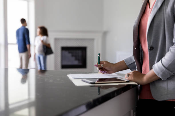 Unrecognizable Real Estate Agent finishes up paperwork Female real estate agent finishes up paperwork as a couple discuss the purchase of a new home in  the background. closing photos stock pictures, royalty-free photos & images