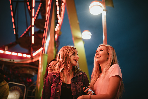 A group of girl friends spend time together at their local county fair carnival, tasting fun foods and trying out an assortment of amusement park rides.  They wait in line to ride the ferris wheel.
