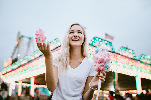 A young woman at her local county fair carnival, tasting fun foods and trying out an assortment of amusement park rides.  She eats some pink cotton candy.