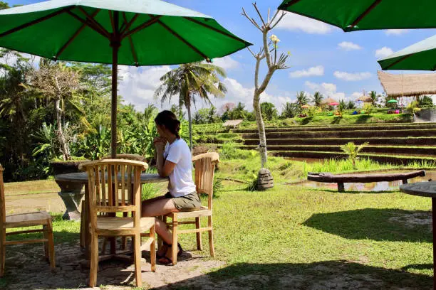 Photo of A beautiful young woman relaxes on a warung's terrace close to typical rice paddies after a trek under the sun in the Campuhan ridge walk, Ubud, Bali.