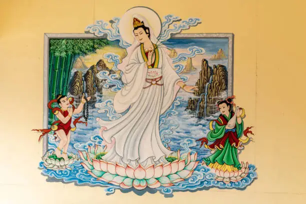 Si Racha, Thailand - March 16, 2019: Closeup of Painting of Guan Yin set against yellow wallin her open circular shrine on Ko Loi Island. Charming lady on lotus floating on water. Devotees present.