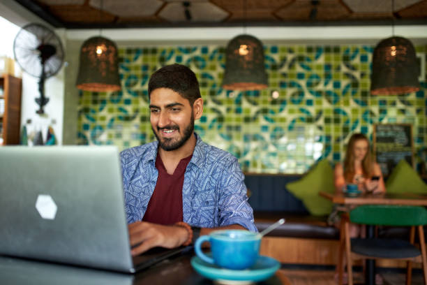 bell'uomo indiano millenario che lavora nel caffè spaziale di coworking alla moda su laptop bevendo cappuccino biologico fair-trade - asian country foto e immagini stock