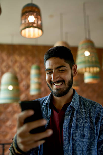 Handsome millennial man with beard using smartphone for video call in organic bohemian cafe Portrait of young attractive, fashionable male texting friends on mobile cellphone in cosy and rustic coffee shop restaurant serving fair-trade pour-over coffee and plant-based food photo messaging stock pictures, royalty-free photos & images