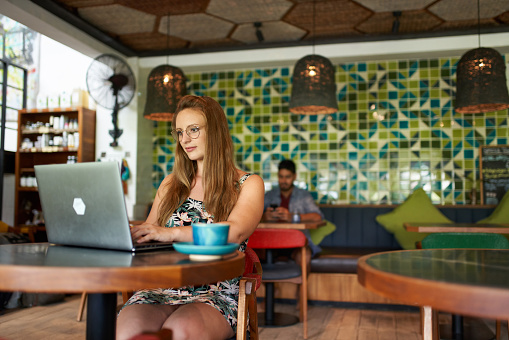 Attractive young female student working on social media marketing with computer in trendy, cosy, tropical organic free-trade coffee shop during travels in Asia