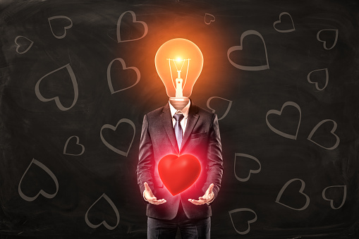 Man in black suit with light bulb instead head, levitating red glaring heart with his palms, standing against black background with heart pattern on it. True love. Genuine feeling. Admire and inspire.