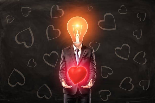 Man in black suit with light bulb instead head, levitating red glaring heart with his palms, standing against black background with heart pattern on it. Man in black suit with light bulb instead head, levitating red glaring heart with his palms, standing against black background with heart pattern on it. True love. Genuine feeling. Admire and inspire. man with a light bulb over his head stock pictures, royalty-free photos & images