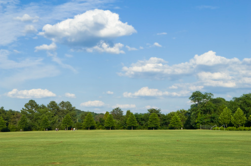 This photo is Soccer Players playing Soccer on Soccer Field with Soccer Ball. the soccer field is lush green grass or lawn or artificial turf at a local park or school. the lighting is natural sunlight. and there are trees, blue sky, white clouds in the background. this photo is a scenic background during the spring or summer. 