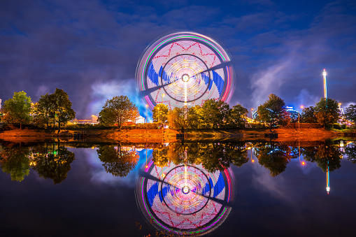 Germany, Illuminated colorful big wheel in motion and rollercoaster of popular folk festival called cannstatter wasen in stuttgart bad canstatt by night reflecting in water