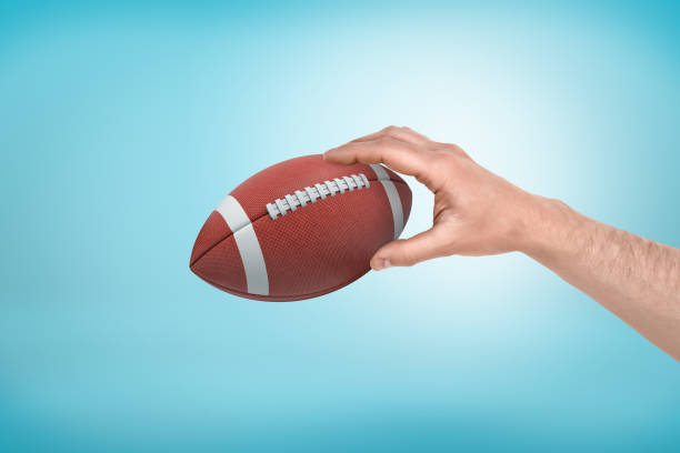 рука человека, держащая мяч для американского футбола на светло-голубом градиенте фона. - football human hand holding american football стоковые фото и изображения