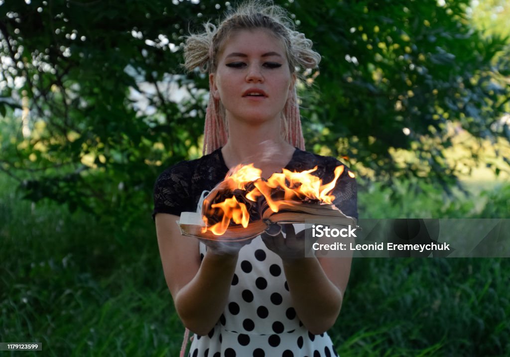 girl holds a burning book in her hands. A young woman in a forest burns a book. The girl holds a burning book in her hands. A young woman in a forest burns a book. Book Stock Photo