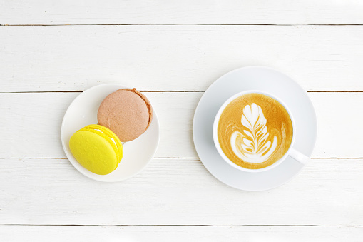 A cup of coffee cappuccino and two macaroons lemon and chocolate on white wooden table. Top view.