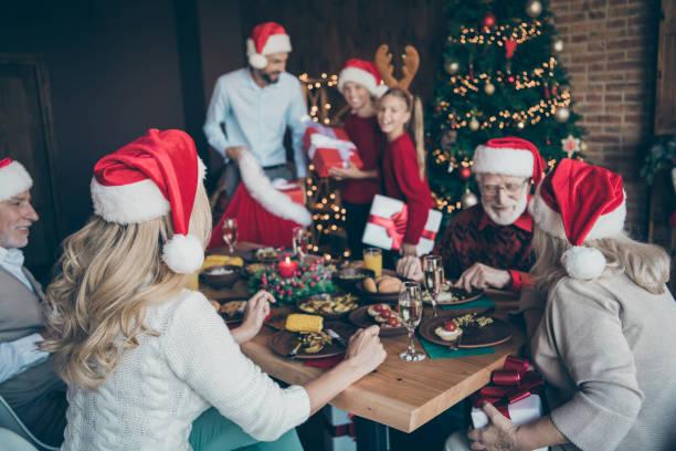 merry x-mas noel meeting of large family back side photo old people sit table speak talk eat feast wear red santa claus cap hat father have fun with small little boy son near christmas tree garlands - senior adult winter senior women daughter imagens e fotografias de stock