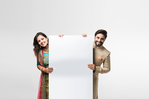 Indian couple holding white board, promoting offers on festival season while wearing traditional cloths, standing isolated over white background