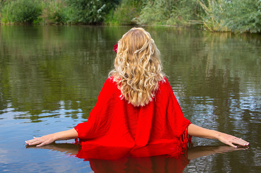 Young blonde woman in red wrap walking in natural water. The young european person is enjoying nature by walking in this dutch lake during summer season. The image is showing the person on her back.