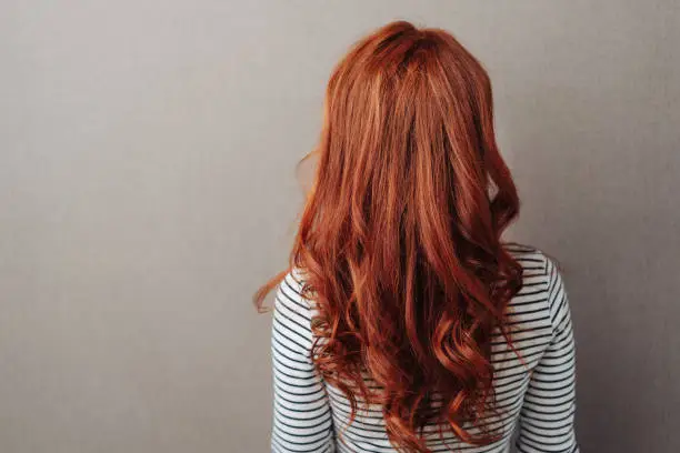 Rear view of a woman with long curly red hair standing facing a grey wall with copy space