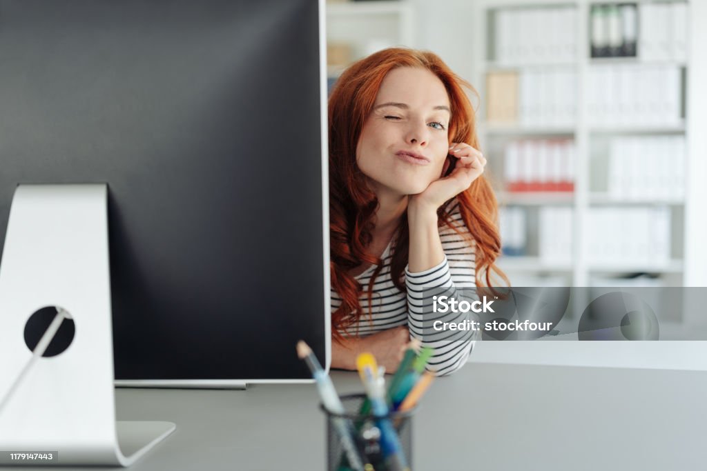 Playful young businesswoman winking at the camera Playful young businesswoman winking at the camera as she peers around the edge of a large desktop monitor in a spacious office Office Stock Photo
