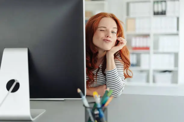 Playful young businesswoman winking at the camera as she peers around the edge of a large desktop monitor in a spacious office