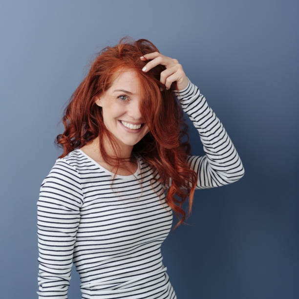 Attractive smiling young woman with long red hair Attractive smiling young woman with long curly red hair standing beaming at the camera with her hand raised to her forehead over a grey background woman hairline stock pictures, royalty-free photos & images