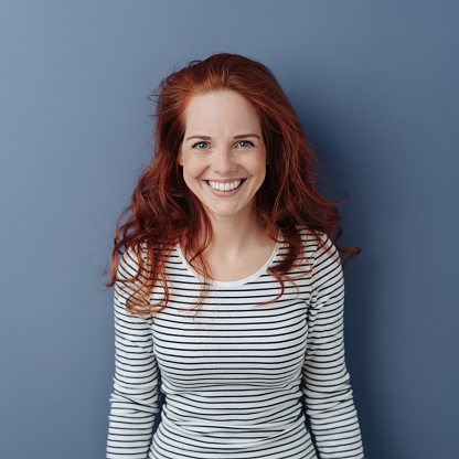 Young redhead woman with a happy pleased smile posing over a grey background looking at the camera with interest