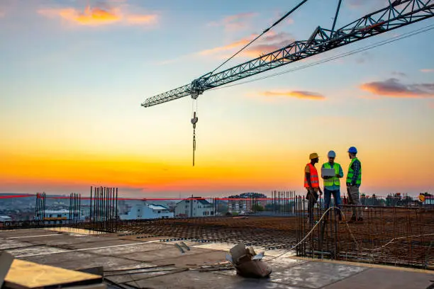Photo of project manager visiting construction site with Multi-Ethnic construction workers standing on roof top at sunset. Business, building, industry, technology and people concept - smiling builder in hardhat with blueprint over group of builders at construction site under the tower crane.