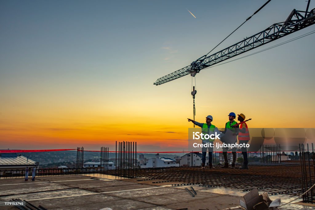 Multi ethic workers talking at construction site reviewing plans Three Multi-Ethnic construction workers in uniform standing at construction site with crane in background, discussing building plans while holding blueprint at sunset under the tower crane. Construction Site Stock Photo