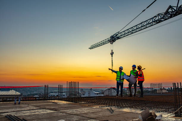 Multi ethic workers talking at construction site reviewing plans Three Multi-Ethnic construction workers in uniform standing at construction site with crane in background, discussing building plans while holding blueprint at sunset under the tower crane. drawing activity photos stock pictures, royalty-free photos & images
