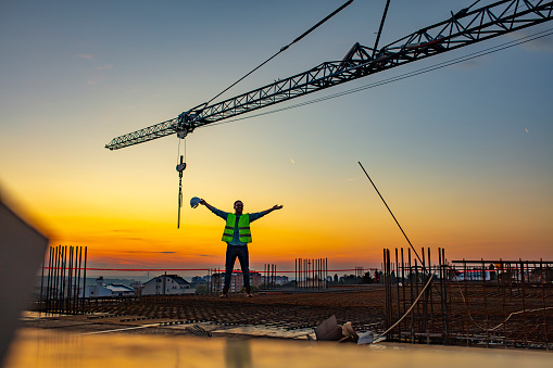 Front view of happy manual worker with arms raised in jubilation standing under tower crane at sunset or sun raise.