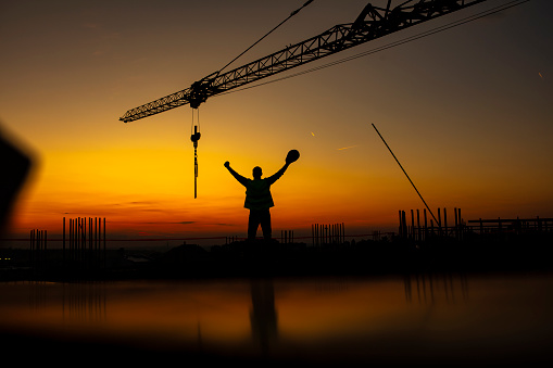 Construction site with crane and Silhouette or young manual worker with arms raised, standing while holding work helmet in his hand at sunset