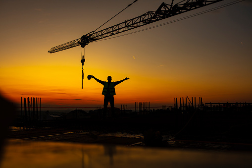 Silhouette of Engineer checking project on building site, construction site at sunset in evening time. Construction engineer with arms up standing under the tower crane.