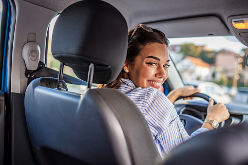 Smiling woman sitting in a car and looking back. Close up portrait of pleasant looking female with glad positive expression, being satisfied with unforgettable journey by car, sits on driver`s seat, enjoys music. People, driving, transport concept