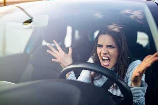 Closeup portrait, angry young sitting woman pissed off by drivers in front of her and gesturing with hands. Road rage traffic jam concept. Woman is driving her car very aggressive and gives gesture with his fist