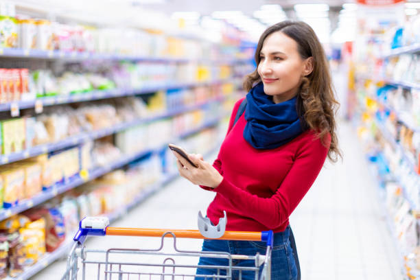 Young smiling woman shopping in a supermarket looking at the screen of the phone her shopping list (grocery list) - consumerism and choosing concept Portrait of woman standing in a supermarket with phone smart phone technology lifestyles chain stock pictures, royalty-free photos & images