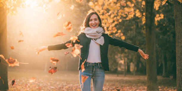 hermosa mujer sonriente con los brazos abiertos y hojas amarillas en el fondo soleado de la naturaleza otoño - autumn women leaf scarf fotografías e imágenes de stock