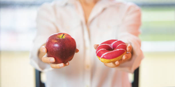 Woman's hands holding red apple and a donut - difficult choice, healthy raw food, sugar and fat addiction concept stock photo
