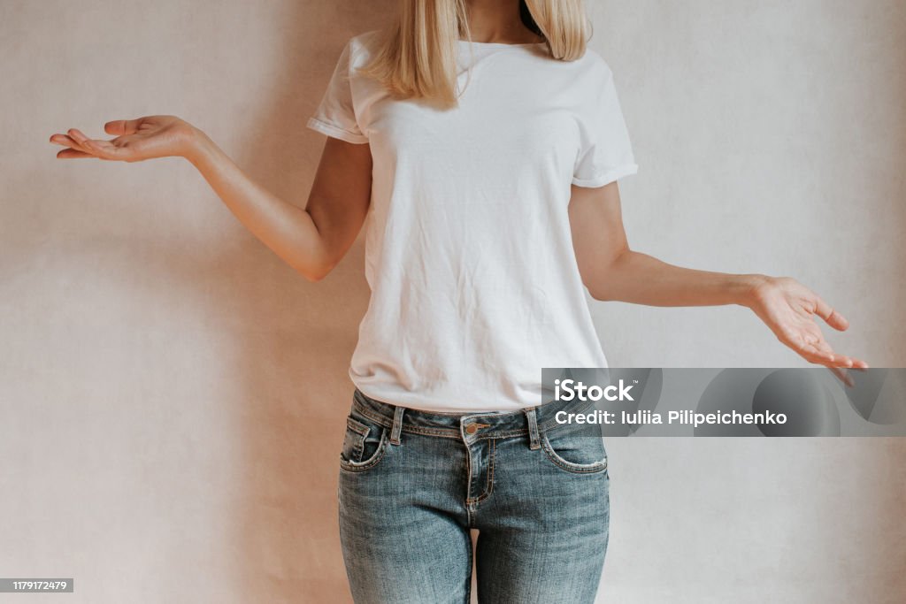 Blond hair woman posing near a light wall. Beautiful young caucasian girl. Hand gestures. Emotion. Casual clothing. Studio model in work. Strong woman, future is female. Jeans and white blank t-shirt T-Shirt Stock Photo