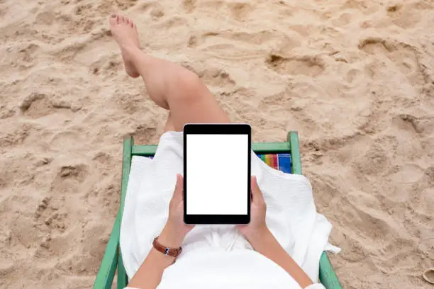 Photo of a woman holding and using a black tablet pc with blank desktop screen while lying down on a beach chair