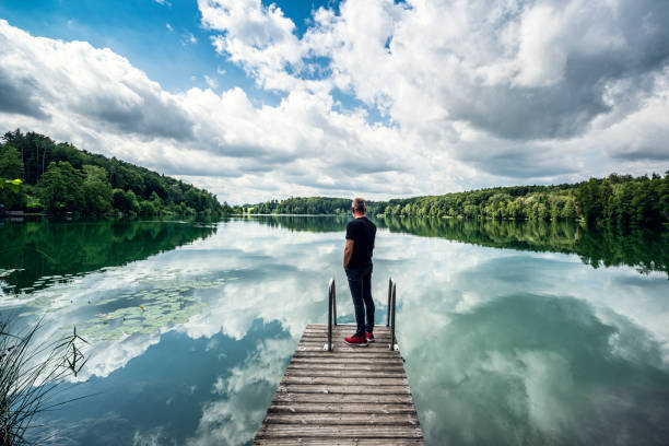 Austria. 40-year-old man with beard stands on a small jetty and looks over the lake.