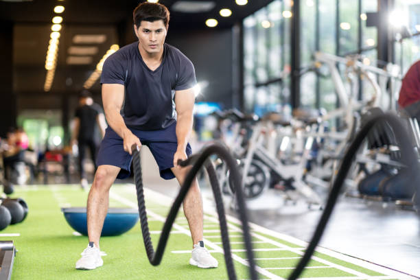 Sporty asian man exercising with battle ropes at the gym on green floor. Strong male determine with her indoor workout for stamina and building muscular body. Athlete battle rope workout concept. Sporty asian man exercising with battle ropes at the gym on green floor. Strong male determine with her indoor workout for stamina and building muscular body. Athlete battle rope workout concept. school gymnasium stock pictures, royalty-free photos & images