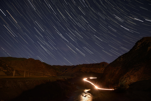 Night landscape with a winding road in the mountains, traces from the headlights of cars and stars in the form of tracks in the sky
