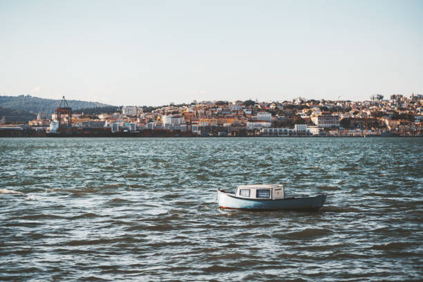An old tiny cozy motorboat with bluish striped curtains sways in the waves of the Tagus river in Portugal, with a riverbank behind full of small antique Lisbon houses and port buildings