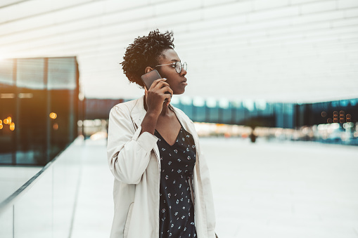 A charming young African woman entrepreneur in a white cloak and spectacles is having a business conversation using a smartphone while standing in an outdoor entrance of a contemporary office building