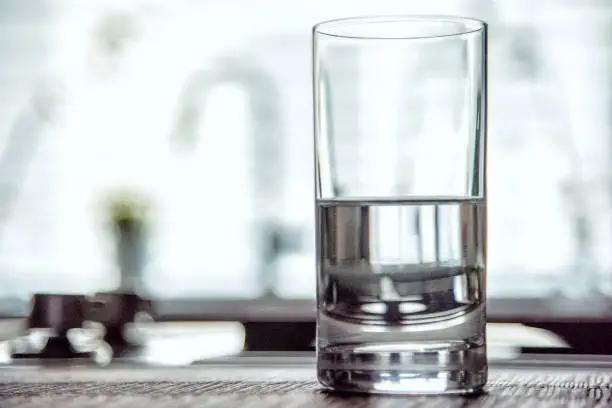 A drinking glass half full of water atop a modern kitchen counter top inside a home as a concept for either optimism (glass half full) or pessimism (glass half empty).