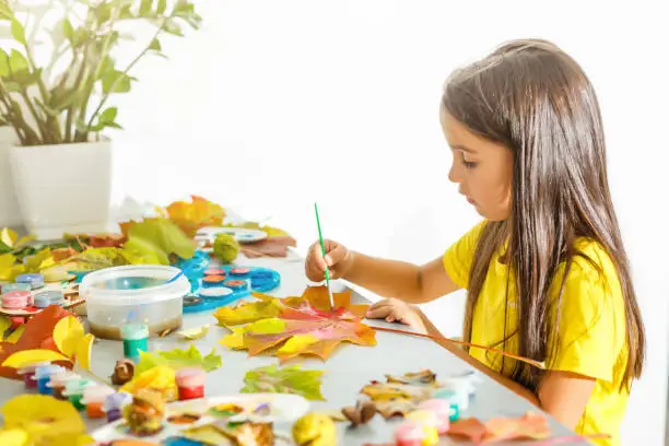 Girl paints leaves. Gouache, brush and various autumn leaves, Children's art project. Colorful Hand-painted on dry autumn leaves