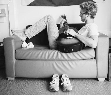 Black and white vintage image style of young man lying on sofa and playing on guitar in teen room.